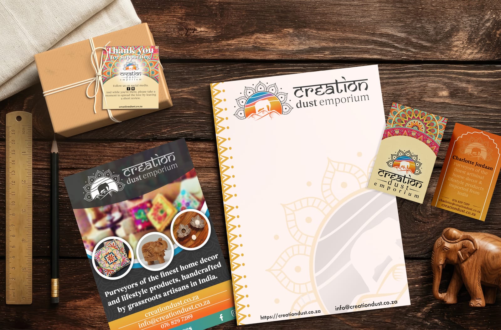 Top-down photograph of Creation Dust Emporium brand collateral including business cards, letterhead, and product flyer on a dark wood background with a carved elephant.
